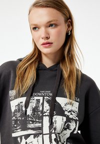 Mørkegrå hoodie med grafiske tryk af bylandskaber og ørne, med en snoretrækshætte og ribbede ærmekanter. Glat, bomuldsagtig tekstur.