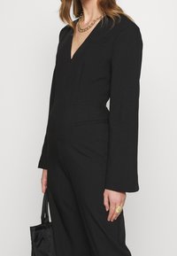 Mono negro con mangas largas, escote en V, forma ajustada y bolsillos laterales. Presenta un cierre con cremallera y tejido suave.