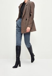 Henkilö, jolla on päällään ruskea herringbone-takkia, musta paita, siniset korkeavyötäröiset housut ja mustat polviin asti ulottuvat korkeat saappaat, seisoo yksinkertaista taustaa vasten.