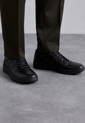 Zapatillas negras con cordones usadas con pantalones oscuros, sobre un suelo de concreto gris.