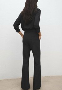 Kobieta o długich ciemnych włosach, ubrana w czarną bluzkę i czarne, rozszerzane spodnie z wysokim stanem, stoi z rękami w kieszeniach na tle gładkiej ściany.