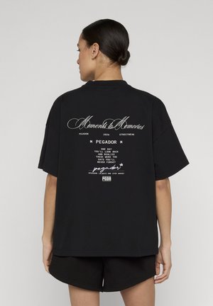 Kvinde med opsat hår iført sort oversized t-shirt med hvid teksten "Moments to Memories" og motiverende tekst på ryggen.
