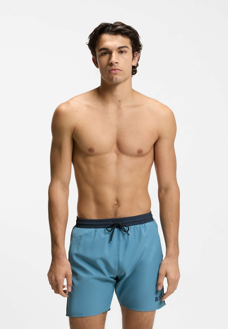 Young man with dark hair wearing light blue swim shorts with black waistband and drawstring, standing against a plain white background.