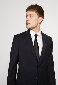 Donker marineblauwe colbert met een enkele knoop, notch revers en opgestikte zakken, gecombineerd met een wit overhemd en een slanke zwarte stropdas. Gladde textuur.