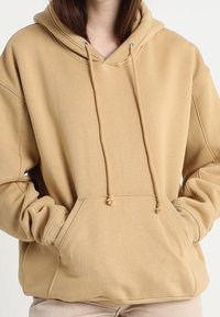 Sudadera beige con un bolsillo tipo canguro en la parte frontal, capucha con cordón y puños acanalados. Tejido suave con una textura lisa. Diseño de corte holgado.