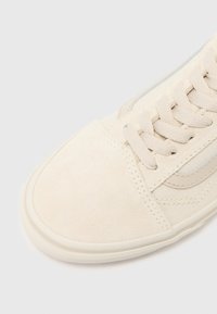 Gros plan sur la pointe d'une basket en daim beige et toile avec des lacets assortis et une semelle en caoutchouc blanche sur un fond clair.