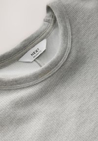 Graues, strukturiertes Rundhalsausschnitt-Shirt mit entspannter Passform, ausgestattet mit einem weichen Stoff und sichtbaren Nähten am Kragen. Das Etikett zeigt den Markennamen.