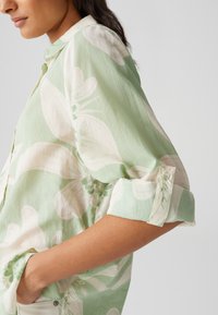 Blusa verde chiaro con grandi motivi floreali bianchi, colletto, maniche arrotolate e una vestibilità comoda con tasche laterali.