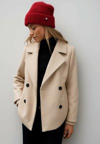 Abrigo beige de doble botonadura con botones negros, solapas anchas y puños acanalados; combinado con un gorro rojo de punto.