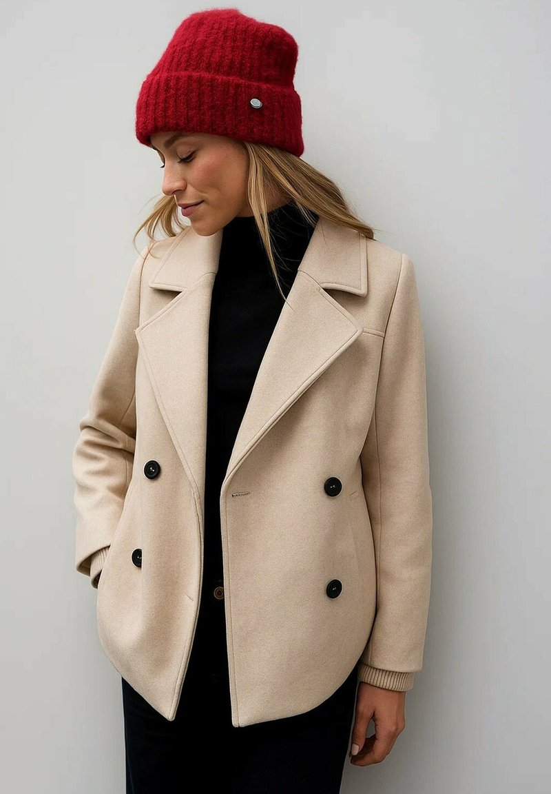 Abrigo beige de doble botonadura con botones negros, solapas anchas y puños acanalados; combinado con un gorro rojo de punto.