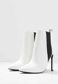 Botines blancos hasta el tobillo hechos de cuero suave con paneles laterales elásticos negros, puntera afilada y un alto tacón de aguja. Diseño limpio con acentos mínimos.