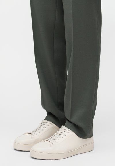 Λευκά δερμάτινα αθλητικά παπούτσια με στρογγυλές μύτες και επίπεδα κορδόνια, σε συνδυασμό με σκούρο πράσινο παντελόνι φαρδιάς γραμμής από λείο ύφασμα.