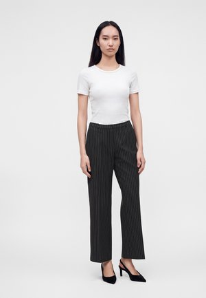 Jeune femme aux longs cheveux foncés portant une chemise blanche à manches courtes, un pantalon large à fines rayures noires et des chaussures à talons noires, debout devant un fond gris.