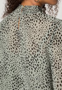 Blusa verde claro con un patrón de manchas negras, con un escote fruncido y una apertura en forma de llave en la parte posterior, terminada con acentos de botón.