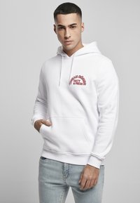 Hvit hettegenser laget av myk bomull, med kengurulomme, snor i hetta, og rød brodert tekst på brystet. Brukt sammen med lyseblå jeans.