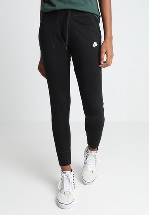 Osoba ubrana w czarne dresowe spodnie Nike i białe wysokie buty sportowe z wzorem w szachownicę na jednolitym tle.