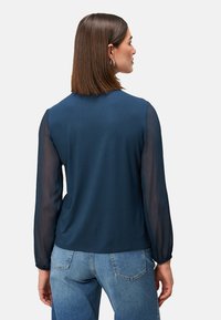 Marineblaues Oberteil mit langen, transparenten Ärmeln und einem einfarbigen Körper. Glatte Textur mit Rundhalsausschnitt. Kombiniert mit hellblauen Jeans.