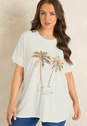 Frau mit langem braunem Haar, die ein weißes T-Shirt mit zwei goldenen Pailletten-Palmen und dem Schriftzug "Palm Springs Beach Club" trägt.