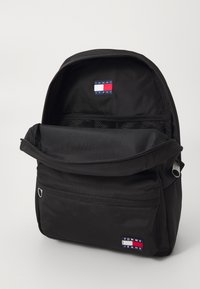 Sac à dos en nylon noir avec une poche zip à l'avant, un compartiment en mesh et un écusson logo cousu en marine, blanc et rouge. Design simple et fonctionnel.