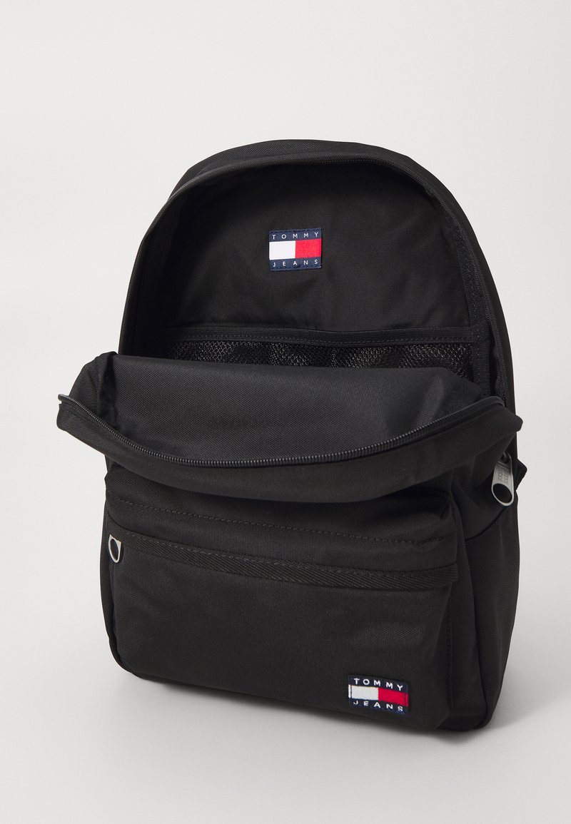 Zaino in nylon nero con una tasca anteriore con zip, scomparto in rete e una patch logo ricamata in blu, bianco e rosso. Design semplice e funzionale.