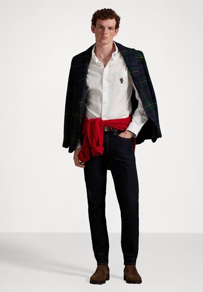 Jauns vīrietis, kuram plecos uzlikts tumši svītrots žakets, virsū balta krekla ar izšūtu lāci, sarkans džemperis saistīts viduklī, tumši džinsi un brūni zābaki.