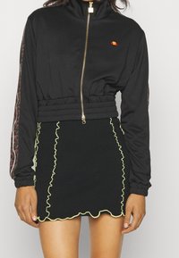 Black zip-up jacket with elastic waist, featuring leopard print sleeves and a small logo. Black skirt with wavy yellow trim and ribbed texture.