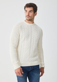 Pulover crem tricotat cu un model de cablu texturat, cu mansete și tiv dintr-o bandă rib, purtat cu blugi albaștri. Dispune de un decolteu rotund și mâneci lungi.
