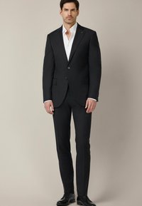 Czarny garnitur o dopasowanym kroju z marynarką na jeden guzik, klapami w kształcie zęba i dwoma guzikami. Biała koszula pod spodem. Spodnie slim-fit. Skórzane buty.