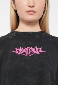 Tricou de mari dimensiuni, de culoare neagră, cu un finisaj texturat. Prezintă un text grafic îndrăzneț de culoare roz "Santa Cruz" în centru, împreună cu elemente decorative.
