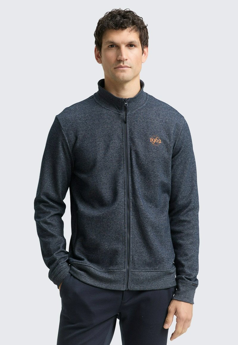 Donkergrijze jas met rits en een hoge kraag, gemaakt van gestructureerde stof, met een oranje logodetail op de borst.