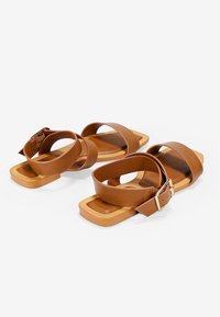Sandalias de cuero marrón con dos correas anchas que cruzan los dedos y una correa trasera asegurada por una hebilla dorada. Suela plana de forma rectangular.