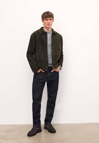 Veste en daim vert olive avec un col, des boutons sur le devant et une poche poitrine, associée à un jean foncé et des chaussures marron sur un fond neutre.