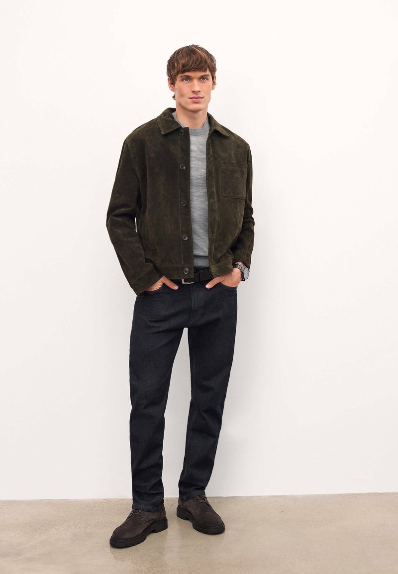 Veste en daim vert olive avec un col, des boutons sur le devant et une poche poitrine, associée à un jean foncé et des chaussures marron sur un fond neutre.