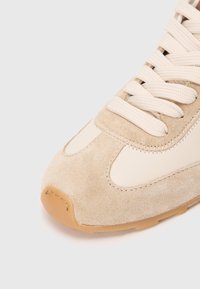 Sneaker bej din suede cu accente de piele crem, având șireturi plate bej și o talpă din cauciuc. Include cusături decorative și un vârf rotunjit.