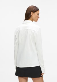 Balta garām piedurknēm krekls ar apkakli, gluda tekstūra un noapaļots apakšmalas dizains. Ikdienišķs stils, kombinēts ar melniem apakšiem. Aizmugures skats.