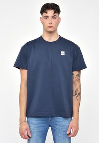 Námorně modré bavlněné tričko s kulatým výstřihem a krátkými rukávy. Obsahuje malou nášivku loga na levé části hrudi. Hladká textura, volný střih.