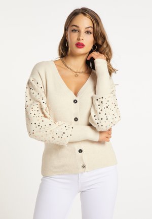 Woman with brown hair wearing cream-colored buttoned cardigan with sequined sleeves and white pants, posing with one hand near her face.