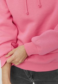 Sweat-shirt en polaire rose vif avec une coupe décontractée, des poignets côtelés et une capuche à cordon de serrage. Comprend un détail logo discret sur la manche.