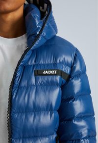 Hornej polovici tela osoby v modrej puffrovacej bunde s čierno-bielou nálepkou "JACK1T" na hrudi a bielou košeľou pod ňou.
