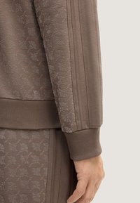 Brauner Textilstoff mit floralem Muster, ausgestattet mit gerippten Bündchen und einem körpernahen Design. Seitliche Streifen sorgen für dezente Details des Kleidungsstücks.
