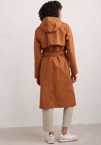 Długi brązowy płaszcz trenchowy wykonany z gładkiego, strukturalnego materiału, z kapturem, z paskiem w talii i detalem na tyle. Nosi się go z jasnymi spodniami i trampkami.
