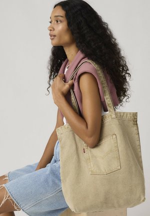 Donna con capelli lunghi e ricci in abbigliamento casual seduta, con una borsa a tracolla in tela beige sulla spalla, davanti a uno sfondo semplice.