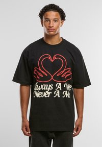 Young man with short curly hair wearing a black oversized T-shirt featuring red swans forming a heart and white text below.