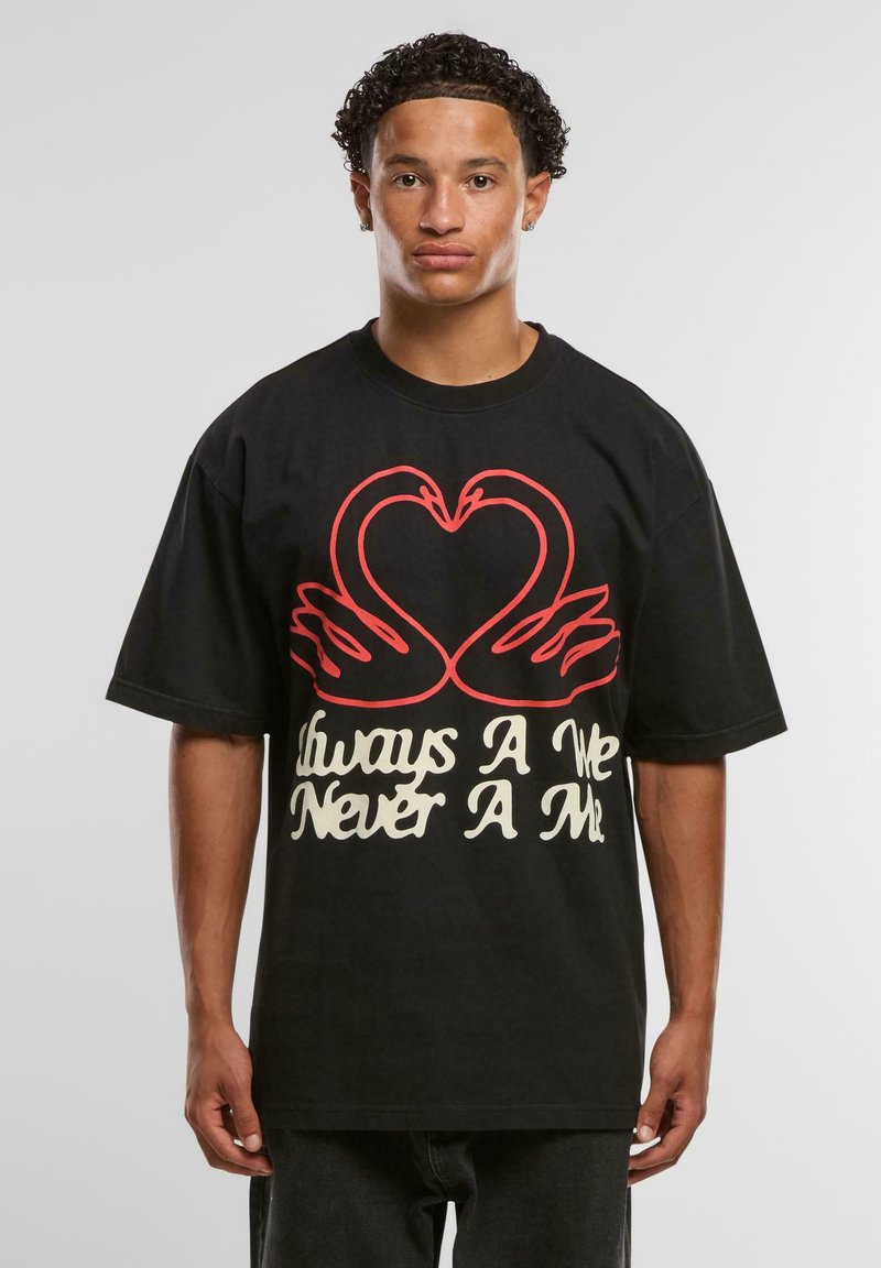 Young man with short curly hair wearing a black oversized T-shirt featuring red swans forming a heart and white text below.