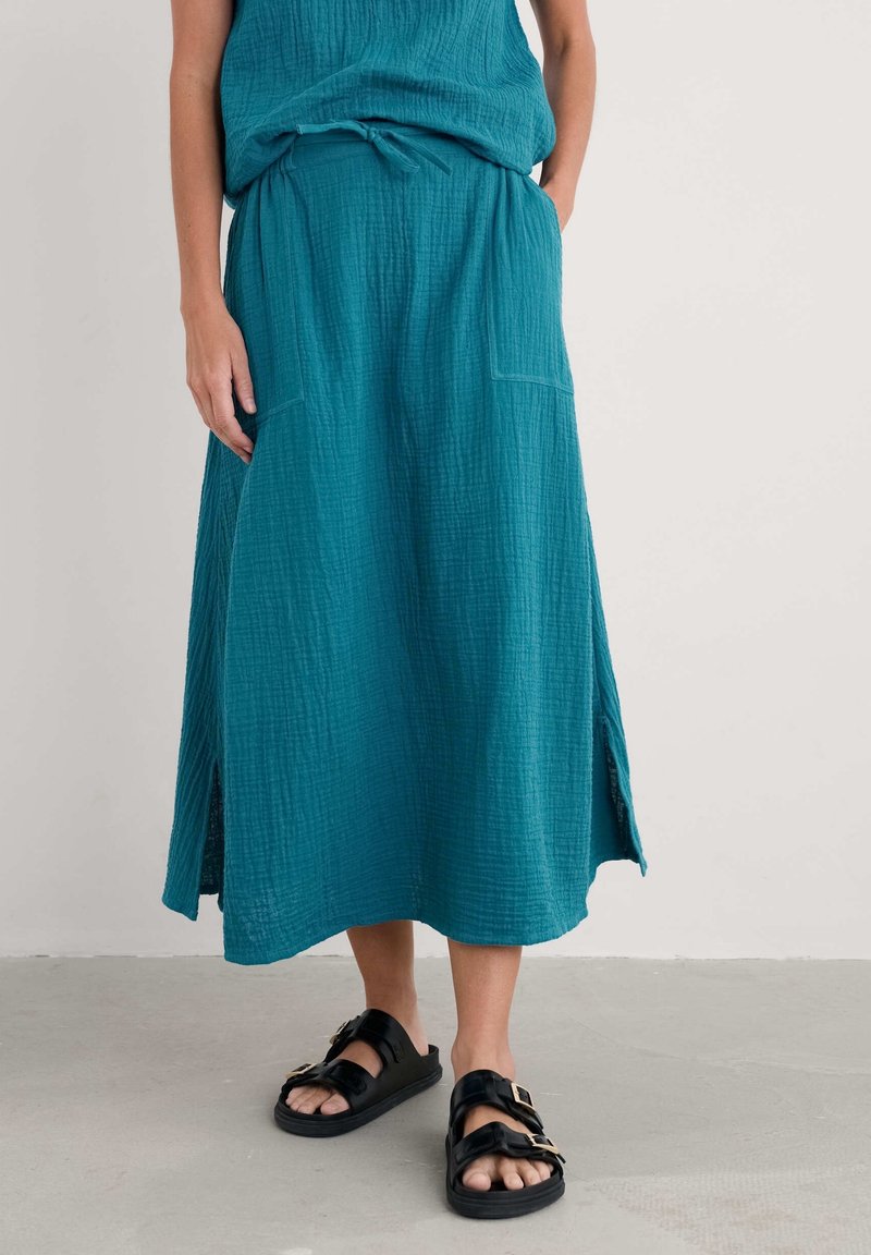 Femme portant une jupe midi texturée bleu sarcelle avec des fentes latérales, des poches et des sandales noires à boucle, debout sur un sol gris contre un mur blanc.