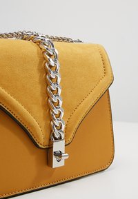 Borsa gialla con patta in suede testurizzato, corpo liscio, tracolla in catena argentata e dettaglio della chiusura rettangolare.