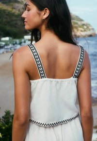 Robe blanche avec des bretelles noires brodées et un dos froncé. Tissu doux et texturé avec une silhouette fluide.