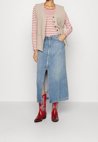 Beige Weste über einem rot-weiß gestreiften Langarmshirt, kombiniert mit einem Jeans-Maxikleid mit einem Reißverschluss vorne und roten Cowboy-Stiefeln mit weißen Akzenten.
