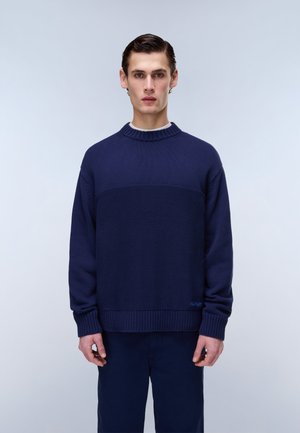 Young man standing still, wearing a dark blue knit sweater and matching pants against a plain light background.