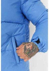 Blauwe pufferjas met een ritszak, elastische manchetten en een gladde, glanzende textuur. Detail van een getatoeëerde hand in de buurt van de zak.